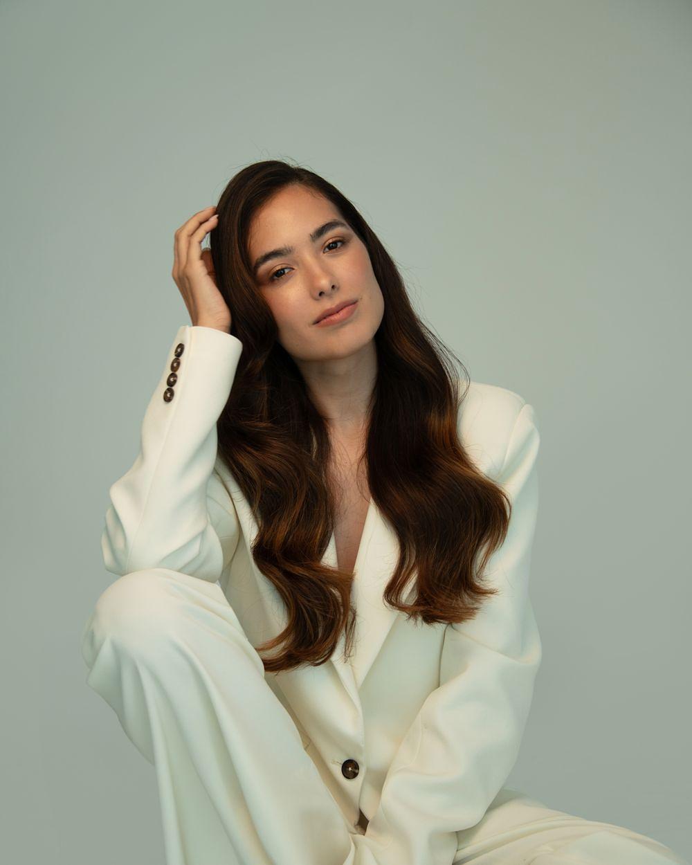 Sharon seated in a cream blazer and matching trousers against a muted green background, with one hand resting in her hair.